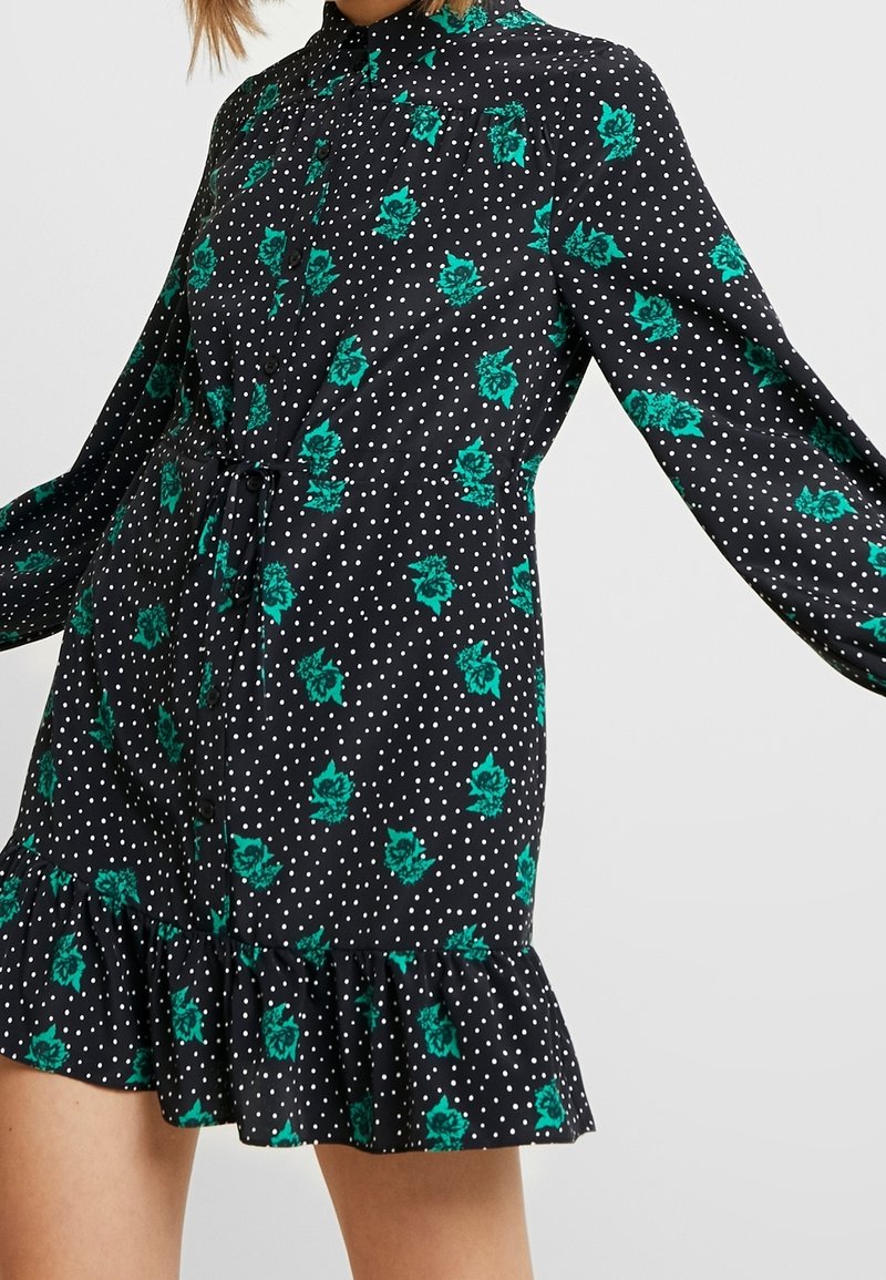 Robe noire à manches longues avec motif floral vert et pois blancs, boutonnée devant, ceinture à la taille et ourlet à volants.