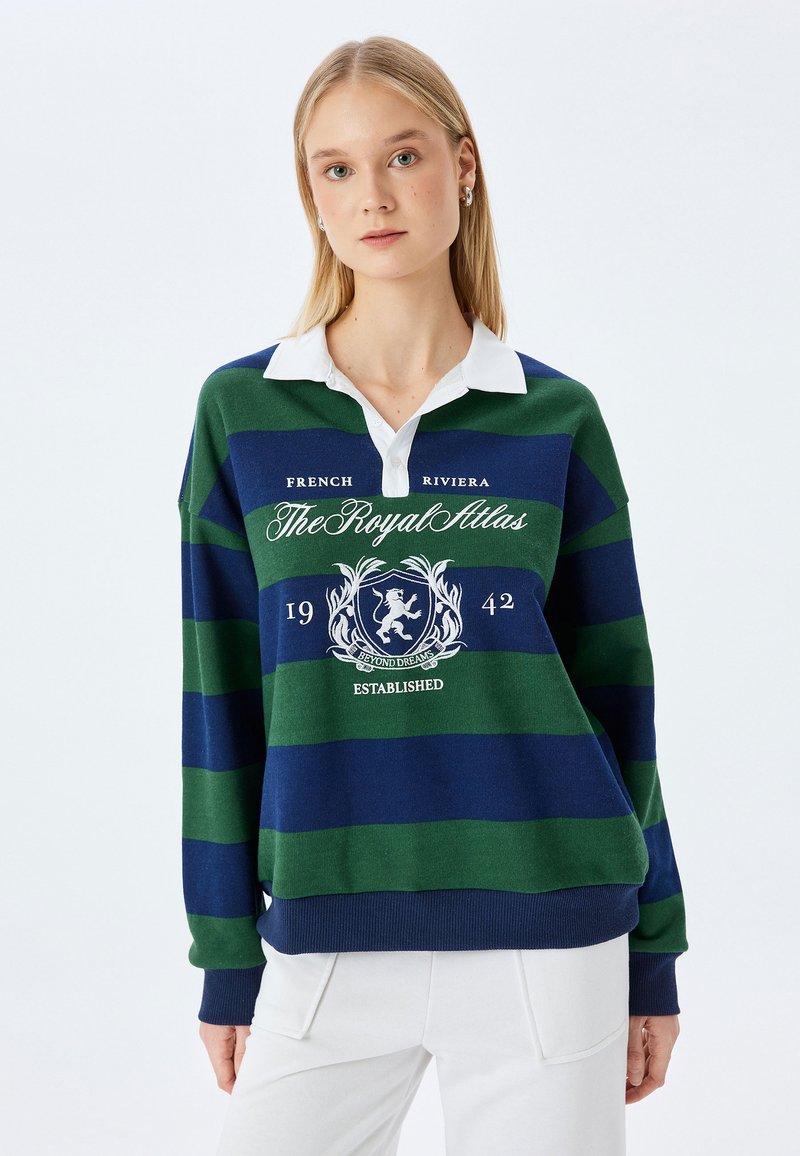 Zielono-granatowy sweter typu rugby w paski z białym kołnierzykiem; zawiera logo i graficzny tekst na klatce piersiowej, a także ściągacze przy mankietach i u dołu.
