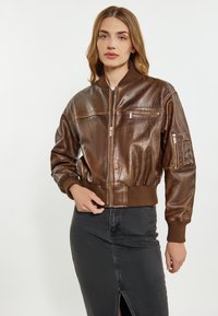 Jachetă bomber din piele maro cu manșete și tiv ribbed, dotată cu buzunare cu fermoar și o textură netedă. Modelul poartă o fustă din denim neagră.