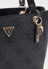 Geantă de mână neagră cu textură, cu accesorii aurii, având un logo triunghiular proeminent Guess și accente decorative în formă de breloc.