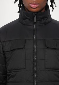 Giacca a vento nera con colletto alto, chiusura con zip e due tasche sul petto, caratterizzata da una texture trapuntata e un tessuto liscio.