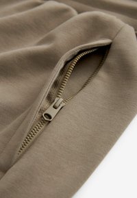 Tissu kaki avec fermeture éclair sur le côté ; texture lisse avec un léger éclat ; zip de couleur assortie, détail de surpiqûres apparentes.