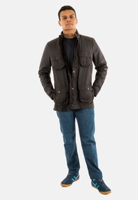 Bruine gewaxte canvas jas met vier voorzakken, een rits aan de voorkant en een kraag. Draagt over een zwart shirt, gecombineerd met spijkerbroek en sneakers.