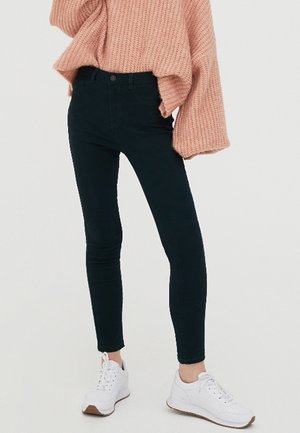 Kvinde iført sorte skinny jeans, en pink chunky striktrøje med brede ærmer og hvide snørebåndssneakers, stående mod en ensfarvet baggrund.
