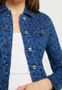 Blue denim jacket featuring a geometric pattern, button closures with "Lee" branding, and a fitted design.