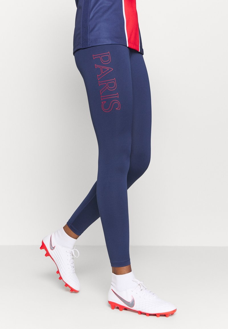 Jambes portant des leggings bleu marine avec "PARIS" en rouge sur la cuisse, des chaussettes de sport blanches et des crampons blancs avec des semelles rouges et un logo swoosh gris.
