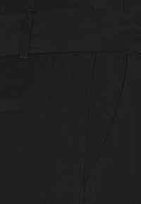 Gros plan sur un pantalon noir sur mesure montrant la taille, les passants de ceinture, la couture latérale et le détail de la poche sur un tissu texturé.