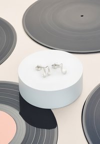 Boucles d'oreilles en forme de notes de musique argentées présentées sur un piédestal circulaire blanc, entourées de divers disques vinyles dans des tons de gris et de rose.