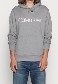 Szara bluza z kapturem wykonana z mieszanki bawełny, z przednią kieszenią, ściągaczami przy mankietach i kapturem ze sznurkiem. Duże białe logo "Calvin Klein" nadrukowane na przodzie.