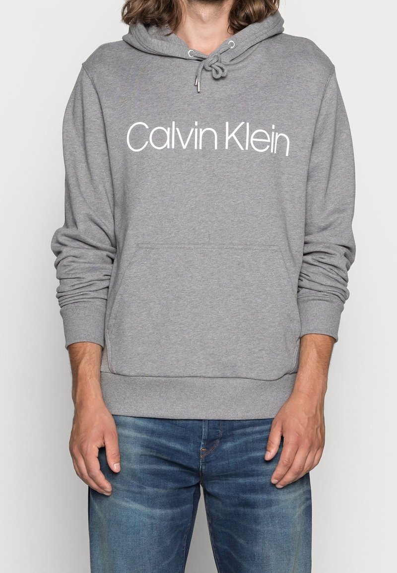 Szara bluza z kapturem wykonana z mieszanki bawełny, z przednią kieszenią, ściągaczami przy mankietach i kapturem ze sznurkiem. Duże białe logo "Calvin Klein" nadrukowane na przodzie.
