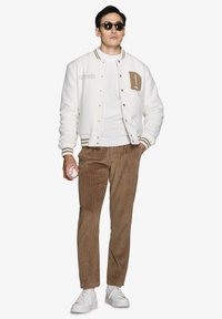 Hvid varsity-jakke med brune accenter, ribbede manchetter og broderede detaljer, parret med brune fløjlspants og hvide sneakers. Holder en baseball.