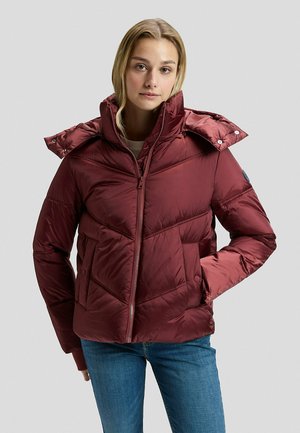 Frau mit glänzender, weinroter Daunenjacke mit Kapuze, blauer Jeans und neutralem Gesichtsausdruck steht vor einem schlichten, hellgrauen Hintergrund.