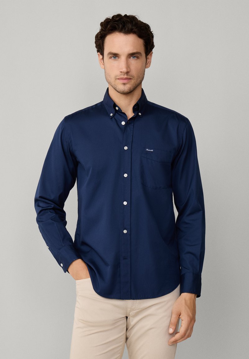 Chemise boutonnée à manches longues bleu marine avec une poche poitrine, en tissu lisse, présentant un logo discret sur le côté gauche.