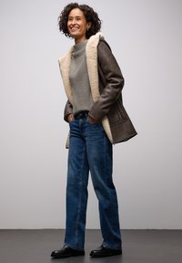 Ruskea nahkatakki, jossa on beige karvavuori, harmaa neulepaita ja siniset suorat farkut. Mustat kengät; kokonaisuus rennon väljä.