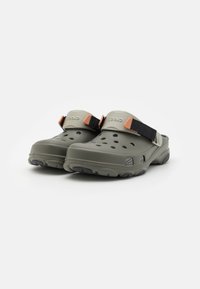 Sabots Crocs verts avec une surface texturée, des trous de ventilation et une sangle noire adjustable avec un accent orange. Semelle en caoutchouc pour une meilleure adhérence.