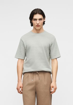 Jeune personne aux cheveux châtains moyens, portant un t-shirt à manches courtes gris clair texturé et un pantalon beige avec cordon de serrage.