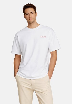 Jonge man met een wit T-shirt met de tekst "AMORE" in roze op de borst, gecombineerd met lichtbeige broeken, handen in de zakken.