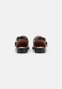 Black patent leather shoes with an open back, featuring a buckle strap and a low, solid heel. Smooth texture and minimalistic design.