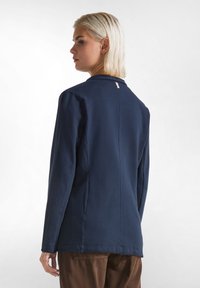 Námorně modrý blazer se strukturovaným designem, dlouhými rukávy a jediným švem na zádech. Obsahuje malou cedulku na výstřihu a hladkou texturu.