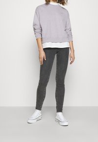 Sudadera de color púrpura claro con cuello redondo y mangas largas, superpuesta a una camisa blanca. Combinada con jeans ajustados grises y zapatillas blancas.