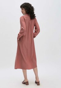 Abito lungo a maniche lunghe di colore rosa chiaro, con colletto, tasche laterali, vestibilità rilassata, dettaglio piega sul retro e lunghezza fino al ginocchio. Indossato con sandali con tacco marroni.
