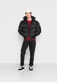 Zwarte pufferjack met ritssluiting en capuchon, gedragen over een rood shirt, gecombineerd met zwarte jeans en zwarte sneakers met witte zolen.