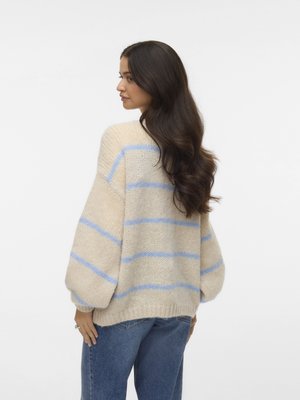 Woman with long dark hair wearing a beige chunky knit sweater with blue stripes and blue jeans, standing against a white background.