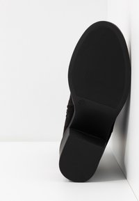 Semelle d'une botte noire avec un talon bas et épais, grip texturé et marquage de taille 37, placée verticale contre un angle blanc.