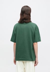 Άτομο με σκούρα μαλλιά μέχρι τους ώμους φοράει ένα χαλαρό σκούρο πράσινο κοντομάνικο πουκάμισο και ανοιχτόχρωμο παντελόνι, στέκεται μπροστά σε ένα απλό φόντο.