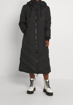 Persona che indossa un lungo cappotto trapuntato nero con cappuccio e cerniera frontale, abbinato a stivali bianchi con lacci e suole spesse nere.