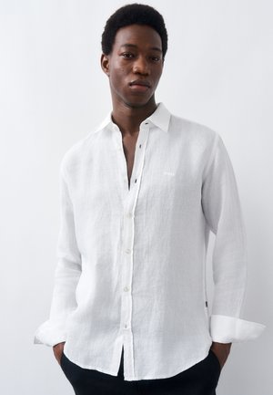 Noor mees seisab käed taskus, seljas valge linane nööpidega särk, taust on üksluiselt valge.