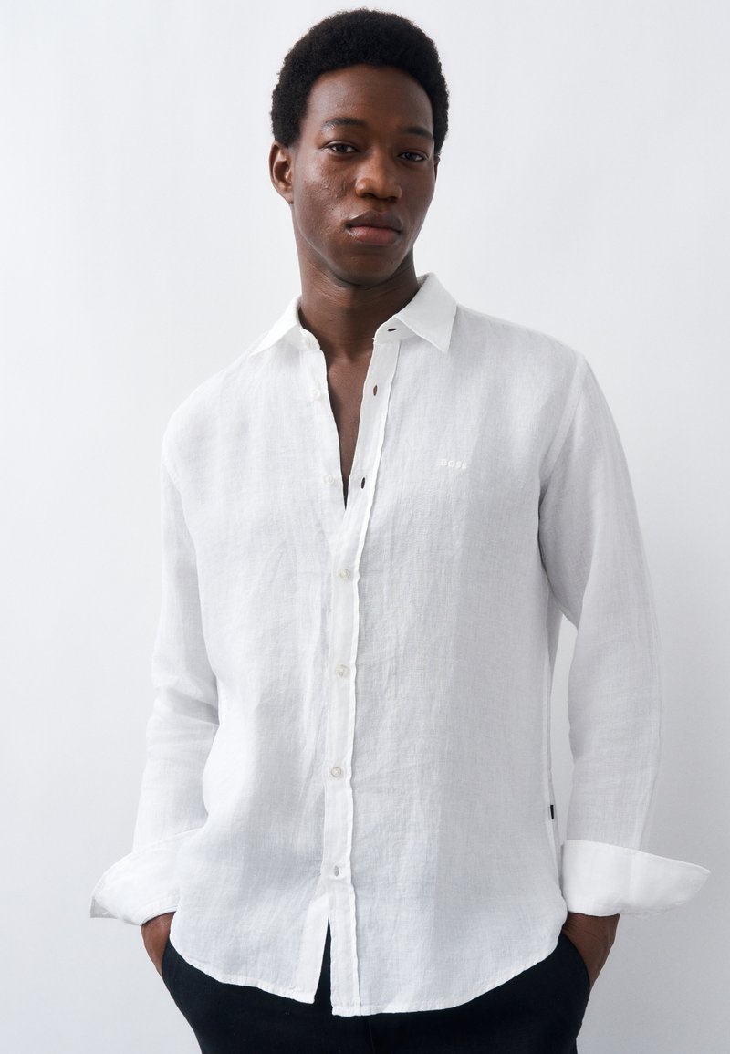 Νέος άνδρας που στέκεται με τα χέρια στις τσέπες, φορώντας λευκό λινό πουκάμισο με κουμπιά, μπροστά σε απλό λευκό φόντο.