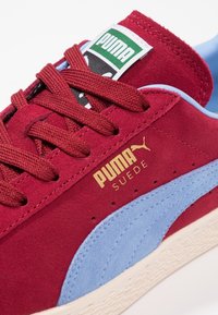 Röd mockaskor med ett blått mockastreck, dekorerade med bruna snören, en gyllene logotyp och en grön och svart märkesetikett på tungan.