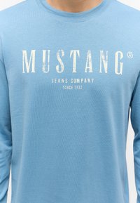 Light blue long-sleeve shirt made of cotton. Features a faded white logo text "MUSTANG JEANS COMPANY SINCE 1932" across the chest.