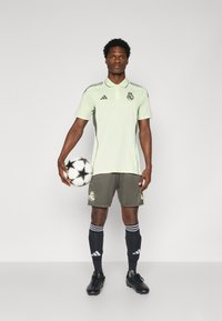 Lichtgroen polo-shirt met zwarte accenten, grijze shorts en zwarte sokken. De speler houdt een gestreepte voetbalschoen vast. Adidas-logo zichtbaar.
