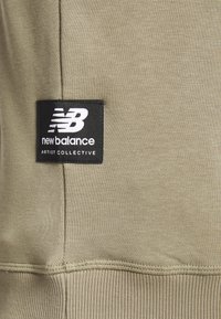 Närbild av beige tyg med en svart New Balance Artist Collective-logotypflicka sydd nära en vertikal söm och ribbad nederkant.