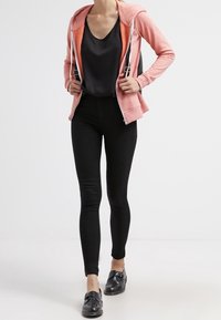 Persona con una sudadera rosa con cremallera, top negro sin mangas, pantalones negros ajustados y zapatos negros sin cordones caminando hacia adelante.