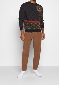 Sort strikket sweatshirt med guldmønstrede accenter og geometriske designs, kombineret med brune fløjlsbukser og hvide sneakers.