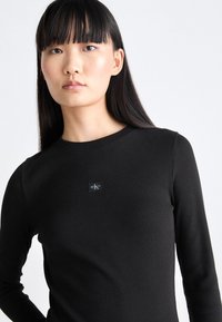 Black ribbed long-sleeve top featuring a round neckline and a small logo patch on the chest. The fabric has a smooth texture.