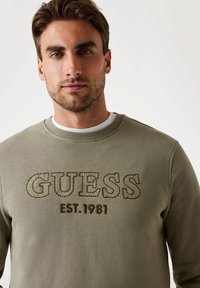 Felpa verde oliva con colletto a girocollo a costine, caratterizzata da un logo testurizzato "GUESS" e "EST. 1981" in marrone. Tessuto morbido, vestibilità casual.