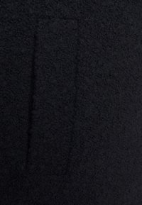 Tessuto nero con una superficie texturizzata e fuzzy. Presenta una toppa rettangolare che potrebbe fungere da tasca; colore solido complessivo senza motivi visibili.