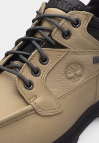 Detailný záber na svetlohnedú koženú topánku s čiernymi šnúrkami, prešívanými detailmi, vyrazeným logom stromu a čiernym vystuženým golierom.