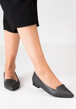 Gros plan de pieds portant des ballerines noires texturées à bout pointu associées à un pantalon noir court sur fond blanc.