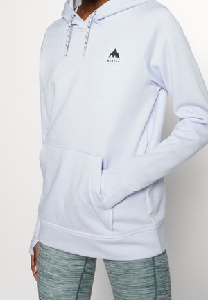 Person wearing a light purple hoodie with a small black mountain logo on the chest and green-gray patterned leggings.