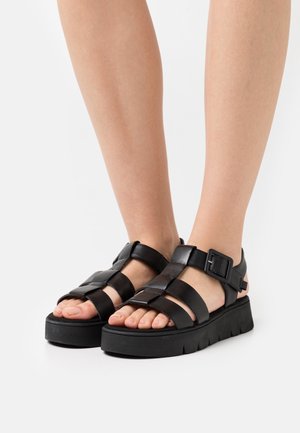 Mustat sandaalit paksulla pohjalla, joissa on leveät ristikkäiset remmit ja soljellinen nilkkaremmi. Valmistettu sileästä nahasta, jossa on matta pinta.