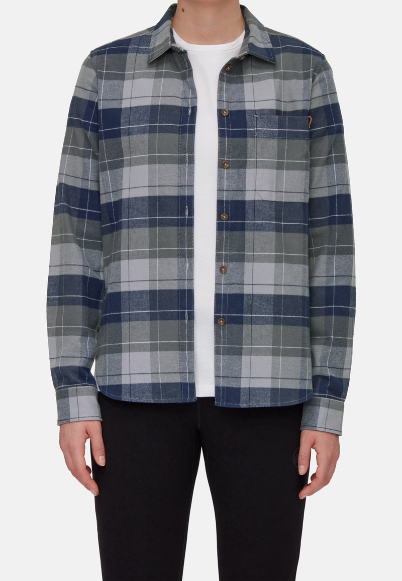 Personne portant une chemise à carreaux bleue, grise et blanche boutonnée sur un t-shirt blanc et un pantalon noir, debout, les bras détendus le long du corps.