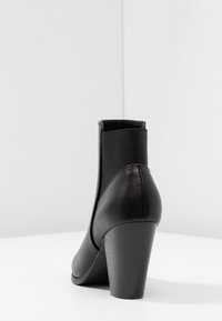 Sort læder ankelstøvle med elastisk sidepanel og stablet blokhæl på hvid overflade mod ensfarvet baggrund.