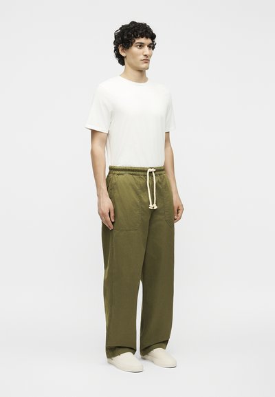 Άνδρας μοντέλο στέκεται, φορώντας ένα λευκό κοντομάνικο μπλουζάκι, φαρδιά παντελόνια ελιάς με κορδόνι και λευκά παπούτσια slip-on.