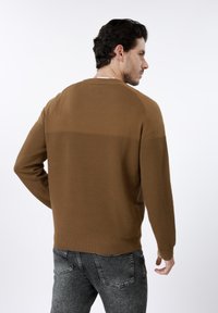 Pull tricoté marron avec texture côtelée, col ras du cou et manches longues. Porté avec un jean gris foncé, présentant un design sans coutures à l'arrière.