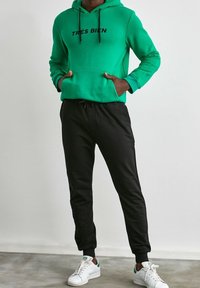 Grön hoodie med texten "TRÈS BIEN", svarta joggers och vita sneakers. Har dragsko, sidofickor och en avslappnad passform.